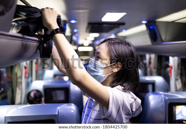 Asian female student stowed her backpack in a\
overhead storage on the school bus,child girl wearing protective\
mask to safety from the Coronavirus,field trip in new normal\
conditions under COVID-19