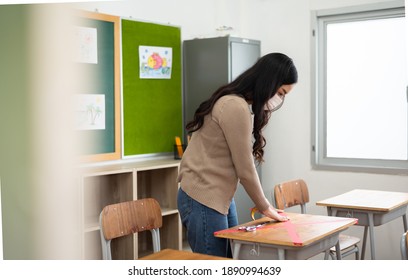 Asian Female Teacher Applying Marking Red Duct Tape In Crossed Symbol On Table In Classroom At School During Virus Pandemic. Young Woman Wearing Protective Face Mask In Social Distancing Concept