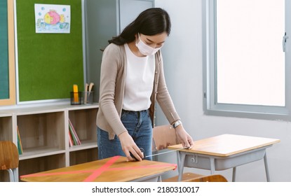 Asian Female Teacher In Face Mask Measuring And Marking Places Distances On Desks In Classroom In Elementary School Before Reopening School. Back To School After Covid-19 Quarantine And Lockdown.