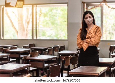 The Asian female teacher is wearing a mask and standing in the empty classroom because of living at risk in the midst of Coronavirus disease 2019 (COVID-19) epidemic. - Powered by Shutterstock