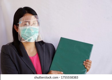 Asian Female Teacher Wearing Protective Facemask And Face Shield , Holding Green Folder Before Going Out To Teach. New Nornal Lifestyle In Covid-19  Ages.