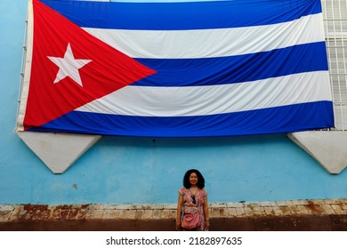 An Asian Female Tourist Standing Under A Large Cuban Flag In Plaza Mayor, Trinidad, Cuba