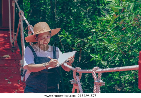 Asian female\
tourist taking notes on suspension bridge while eco tourism in\
mangrove forest at natural\
parkland