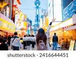 Asian female tourist Traveling and having fun. And she was taking photo with a cell phone camera at Night street with many restaurant around Tsutenkaku Tower in Shinsekai district of Osaka, Japan.