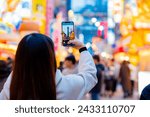 Asian female tourist Traveling and having fun. And she was taking photo with a cell phone camera at Night street with many restaurant around Tsutenkaku Tower in Shinsekai district of Osaka, Japan.