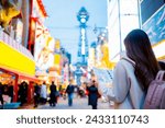 Asian female tourist Traveling and having fun. And she was taking photo with a cell phone camera at Night street with many restaurant around Tsutenkaku Tower in Shinsekai district of Osaka, Japan.