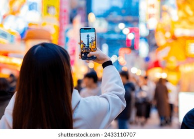 Asian female tourist Traveling and having fun. And she was taking photo with a cell phone camera at Night street with many restaurant around Tsutenkaku Tower in Shinsekai district of Osaka, Japan. - Powered by Shutterstock