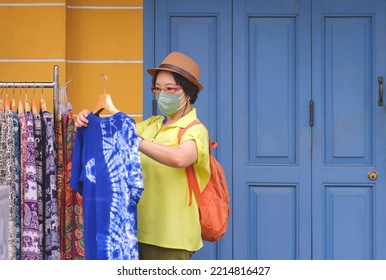 Asian Female Tourist In Vintage Style And Protective Face Mask Choosing Tie Dye T-shirt On Cloth Rack Of Street Clothing Store In Outdoor Market At Bangkok, Thailand