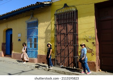 Asian female tourists are standing, posing for a picture in front of the rundown facade of a colonial style building with large windows in Trinidad, Cuba.