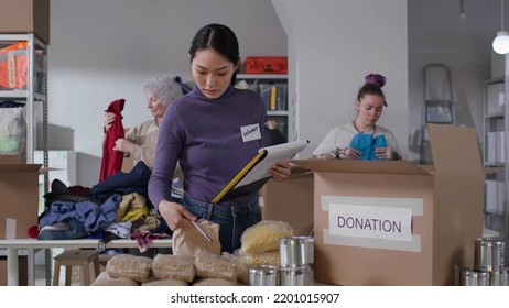Asian female volunteer with clipboard packing food and drinks in boxes at distribution or refugee assistance center - Shutterstock ID 2201015907