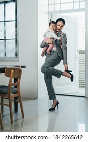 Asian Female Worker Just Reaching Home After Picking Up Her Baby From Day Care Center Is Taking Off Her Heels. Korean Mother Holding Her Baby Is Standing Single Legged , Trying To Put Shoes Off.