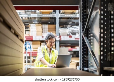 Asian Female Worker In Safety Vest Sitting And Working With Computer Laptop In Storage Warehouse. People, Warehouse And Industry Concept