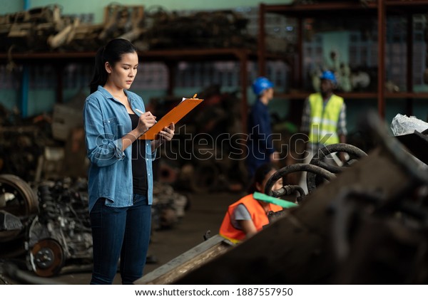 Asian\
female worker working with clipboard and checking old automotive\
spare parts, engine, motor, machine for repair or maintenance in\
garage or automotive spare parts storage\
warehouse