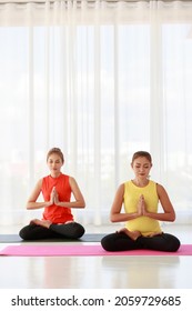 Asian females closing eyes and clasping hands while meditating in Lotus pose during yoga session in studio