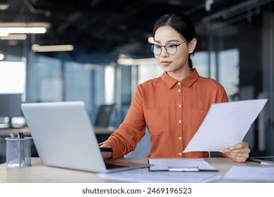 Asian financial woman working inside office with documents, papers behind paperwork, serious businesswoman looking financial reports at workplace , confident successful female accountant. - Powered by Shutterstock