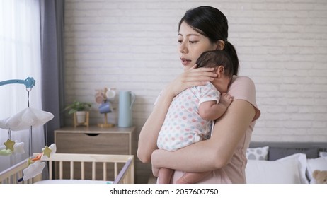 Asian First Time Mom Is Holding Her Cute Little Baby Upright By Supporting Its Neck With Her Hand In The Bedroom With White Wall At Background.