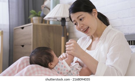 Asian First Time Mom Is Sitting And Holding Tiny Hands With A Smile While Singing Nursery Song To Her Baby Daughter On The Bed In The Bedchamber.