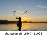 Asian fisherman on wooden boat casting a net for catching freshwater fish in nature river in the early morning before sunrise