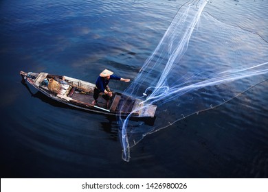 Asian Fisherman On Wooden Boat Casting A Net For Catching Freshwater Fish In Nature River In The Early Morning Before Sunrise