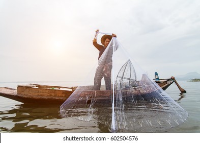 Asian Fisherman On Wooden Boat Casting A Net For Catching Freshwater Fish In Nature River In The Early Morning Before Sunrise