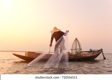 Asian Fisherman On Wooden Boat Casting A Net For Catching Freshwater Fish In Nature River In The Early Morning Before Sunrise
