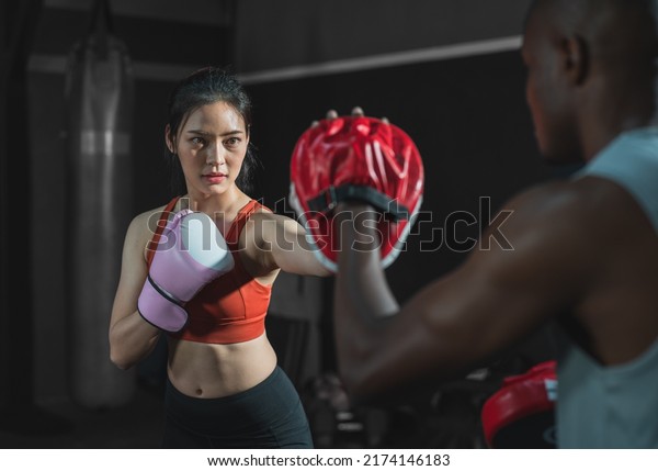 Asian fit female boxing training with her trainer\
at gym.Sportwoman  wearing boxing gloves exercise and punch to pads\
for boxing