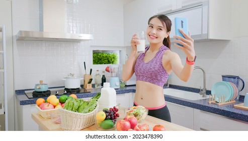 Asian Fitness Woman Holding Milk And Taking Selfie While Standing In Kitchen At Home With Healthy Food