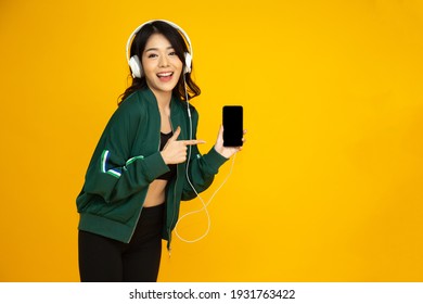 Asian Fitness Woman Listening Music With Headphone On Smartphone And Pointing Finger To Phone Isolated On Yellow Background