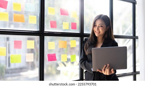 Asian freelance woman smiling paste post-it on the wall to record for work and using on laptop on wooden table at home. Entrepreneur woman working for her business at home. Business concept. - Shutterstock ID 2255793701