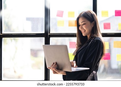 Asian freelance woman smiling paste post-it on the wall to record for work and using on laptop on wooden table at home. Entrepreneur woman working for her business at home. Business concept. - Shutterstock ID 2255793733