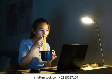 Asian freelancer smart business woman eating instant noodles while working on a laptop in the living room at home at night happy Asian girl sitting on a desk overtime enjoying relaxing time