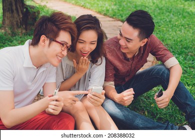 Asian Friends Sitting On The Ground And Sharing Something Funny In The Phone