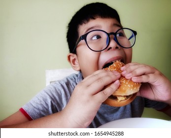 Asian Funny Boy Wearing Glasses, Eating A Delicious Hamburger. Opening His Mouth Wide To Eat. He Likes To Eat Junk Food And Is Happy Every Time. Eating A Lot Causing Health Problems Including Obesity.