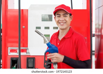 Asian Gas Station Attendant In Red Uniform Stand Smiling, Holding Gas Pump Nozzle At The Gas Station. Cash, Price And Volume Readout Display On Petrol Pump Display Screen.