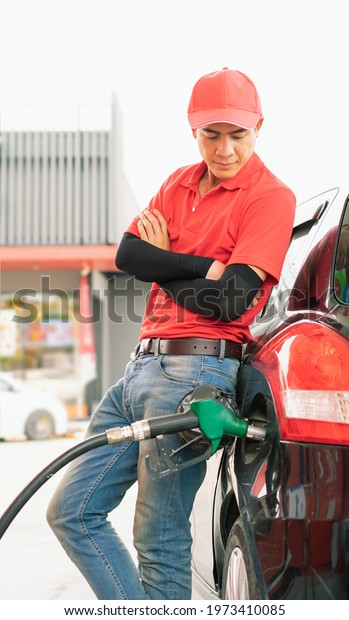 Asian gas station worker leaning against car\
and looking to green fuel nozzle which fill energy fuel into black\
car tank in petrol station, commercial service for benzine, diesel,\
gasohol, gasoline.