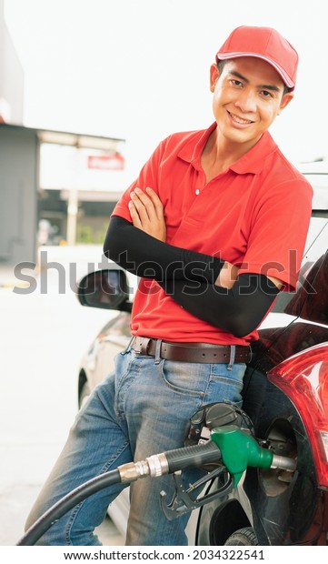 Asian gas station worker man leaning against\
black car and looking straight with smile while green fuel nozzle\
filling high energy power fuel into black auto car tank, commercial\
service for benzine