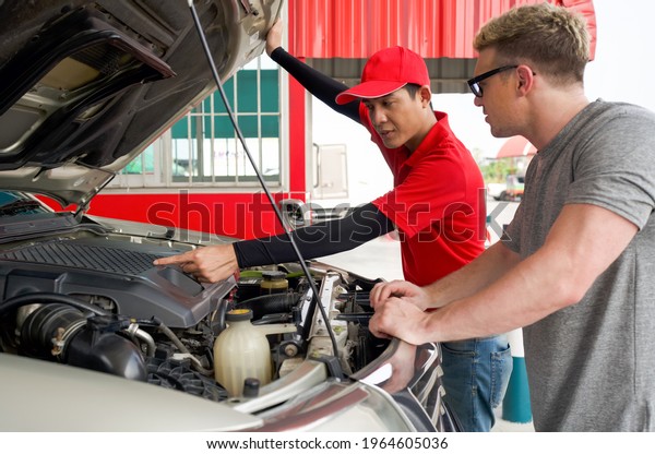 An Asian gas station worker raises the car bonnet\
to check for the cause of an engine malfunction. A caucasian driver\
with sunglass stand listening attentively in front of broken pickup\
truck.