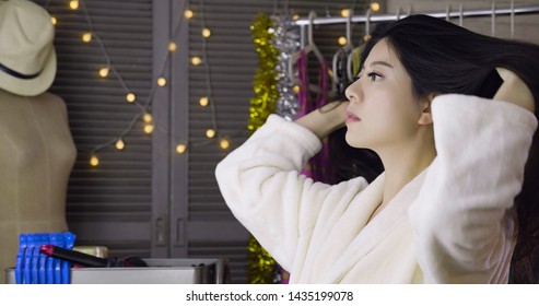 Asian Girl Actress Touching Her Long Black Hair Looking In Mirror In Makeup Room Backstage. Mannequin Dummy Standing Beside Dressing Table With Lights Hanging On Wall. Woman Singer In White Bathrobe