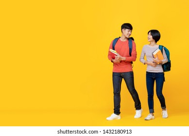 Asian Girl And Boy Students With Bags Holding Books In A Conversation While Walking Towards Class College Isolated Against Yellow Studio Background