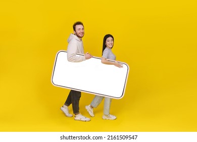 Asian Girl And Caucasian Guy Holding Huge Smartphone Showing Blank White Screen, Mobile App Advertisement And Excited Smile On Camera Isolated On Yellow Background. Product Placement.