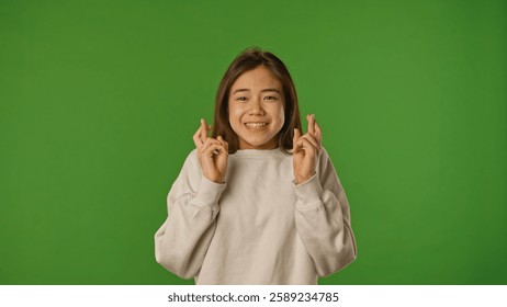 Asian girl child with crossed fingers and please face looking at camera, positive expression, isolated on chroma key green screen studio background mockup.