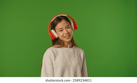 Asian girl child listening music in headphones, positive expression, isolated on chroma key green screen studio background mockup.
