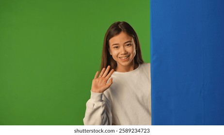 Asian girl child peeks out and waves hand, blue space, positive expression, isolated on chroma key green screen background mockup.