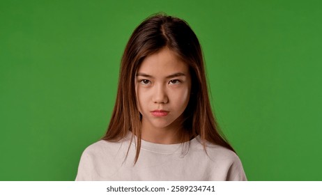 Asian girl child with sad angry face looks at camera, negative expression, isolated on chroma key green screen studio background mockup.