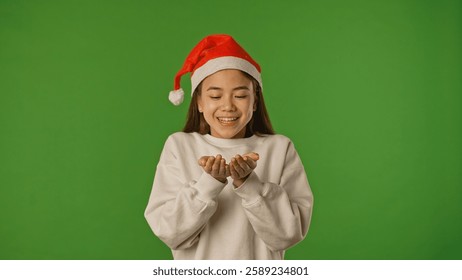 Asian girl child in santa hat blows snow from hands and looks around, positive expression, isolated on chroma key green screen studio background mockup.
