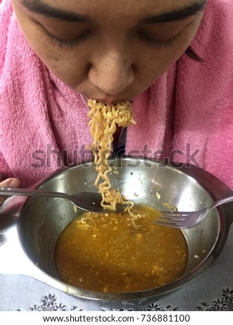 An Asian girl covered up in a pink towel hungrily eating instant noodle soup from a bowl by using a spoon and fork.