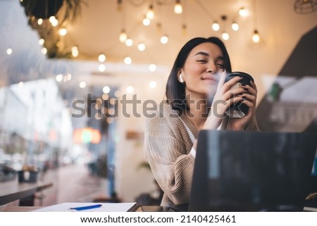 Asian girl drinking and enjoying coffee during working on laptop in cafe. Young millennial brunette woman wearing earphones sitting at table. Freelance and remote work. Modern female lifestyle