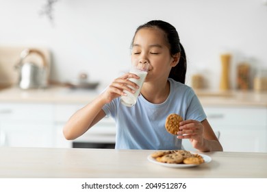 Asian Girl Drinking Milk And Enjoying Cookies, Sitting In Kitchen And Eating Snack, Free Space. Pretty Child Having A Bite With Freshly Baked Biscuits And Calcium Drink