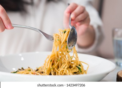 Asian Girl Eating A Plate Of Tasty Italian Spaghetti With Parsley And Herb Garnished On Top Dish In Modern Restaurant. Hands Twirl Spoon Fork To Roll Up Spaghetti As Italian Etiquette. Glass Of Water