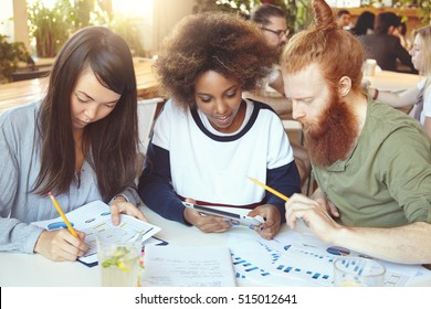 Asian girl filling in papers with graphs and diagrams while African woman sharing ideas with redhead bearded colleague on touch pad. Young businesspeople working on start-up project using tablet - Powered by Shutterstock